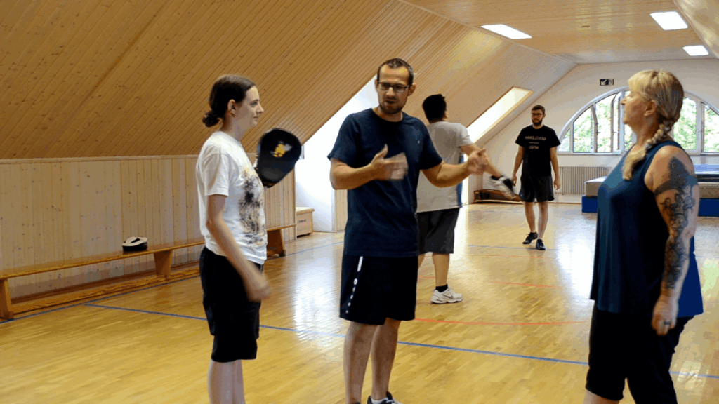 Trainer erläutert eine Partnerübung neben einer Teilnehmerin mit Handpratze